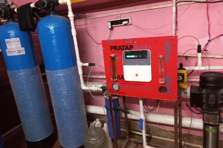 A photo of a red control panel in a room filled with electrical equipment. The panel has several gauges, buttons, and levers for wholesale water supply. Next to the panel are two blue tanks and a water pump.